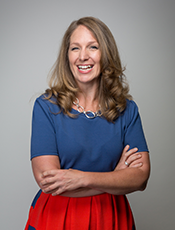 Photo of Janikke Klem from waist up, Caucasian female, long curled dark blonde hair, wearing blue short sleeve blouse tucked into red skirt and ring linked necklace